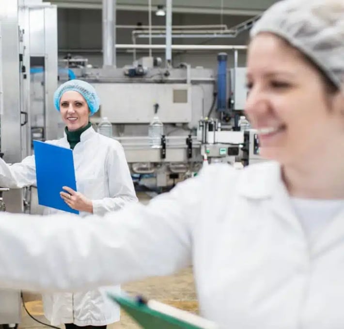Two women in lab coats and hairnets work in a factory or laboratory setting, one holding a blue clipboard and the other a green clipboard, ensuring Food & Beverage Compliance.
