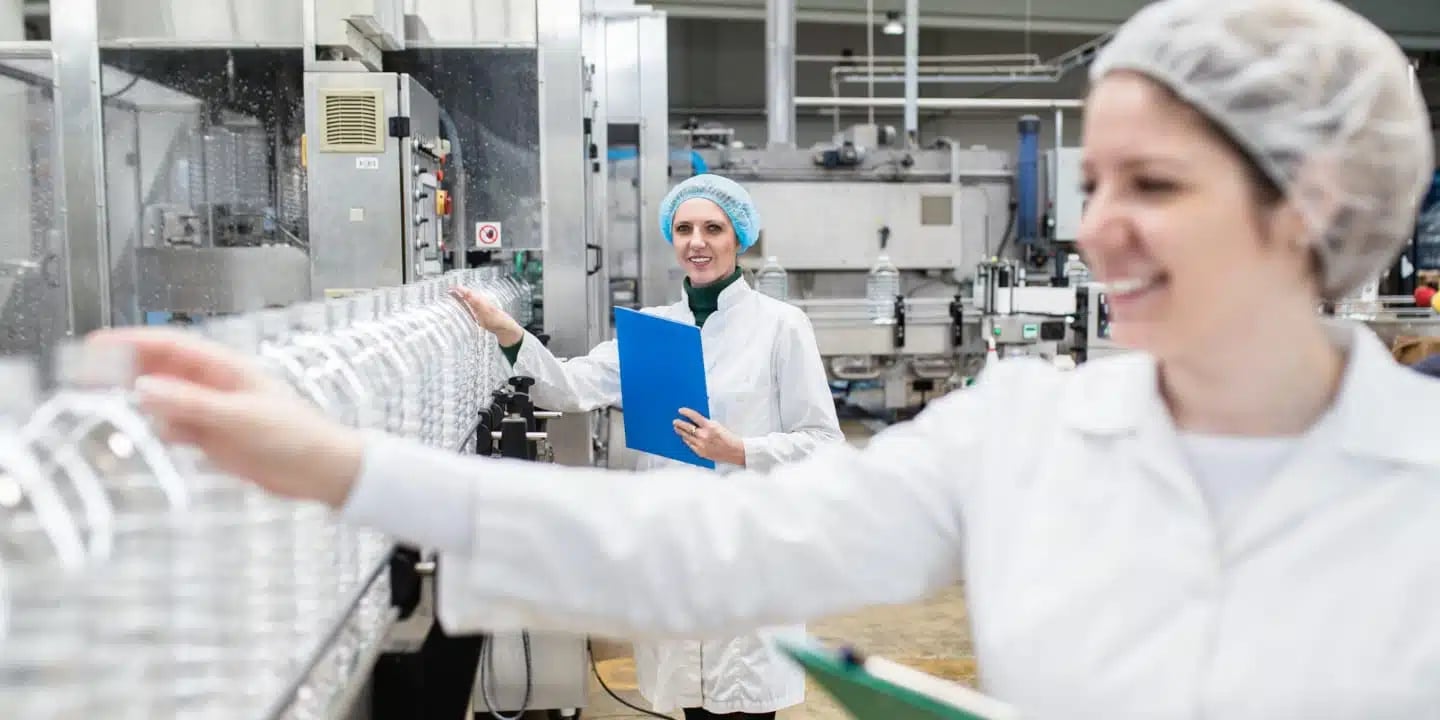 Zwei Mitarbeiter in Laborkitteln und Haarnetzen prüfen Plastikflaschen auf einem Förderband und stellen so die Einhaltung der Lebensmittel- und Getränkvorschriften in einer Fabrikumgebung sicher.