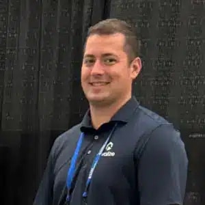 A man in a navy blue collared shirt stands in front of a black curtain, smiling at the camera. He is wearing a conference badge and lanyard.