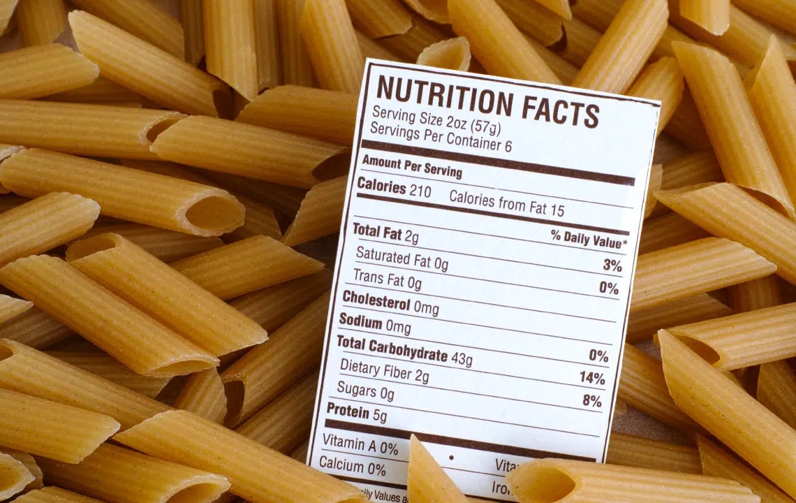 Close-up of uncooked penne pasta with a nutrition facts label showing calories, fat, sodium, carbohydrates, fiber, and protein per serving.
