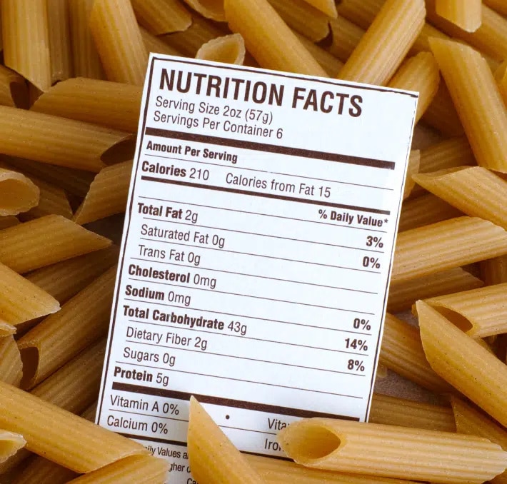 A nutrition facts label displaying information for pasta sits on top of uncooked penne noodles.