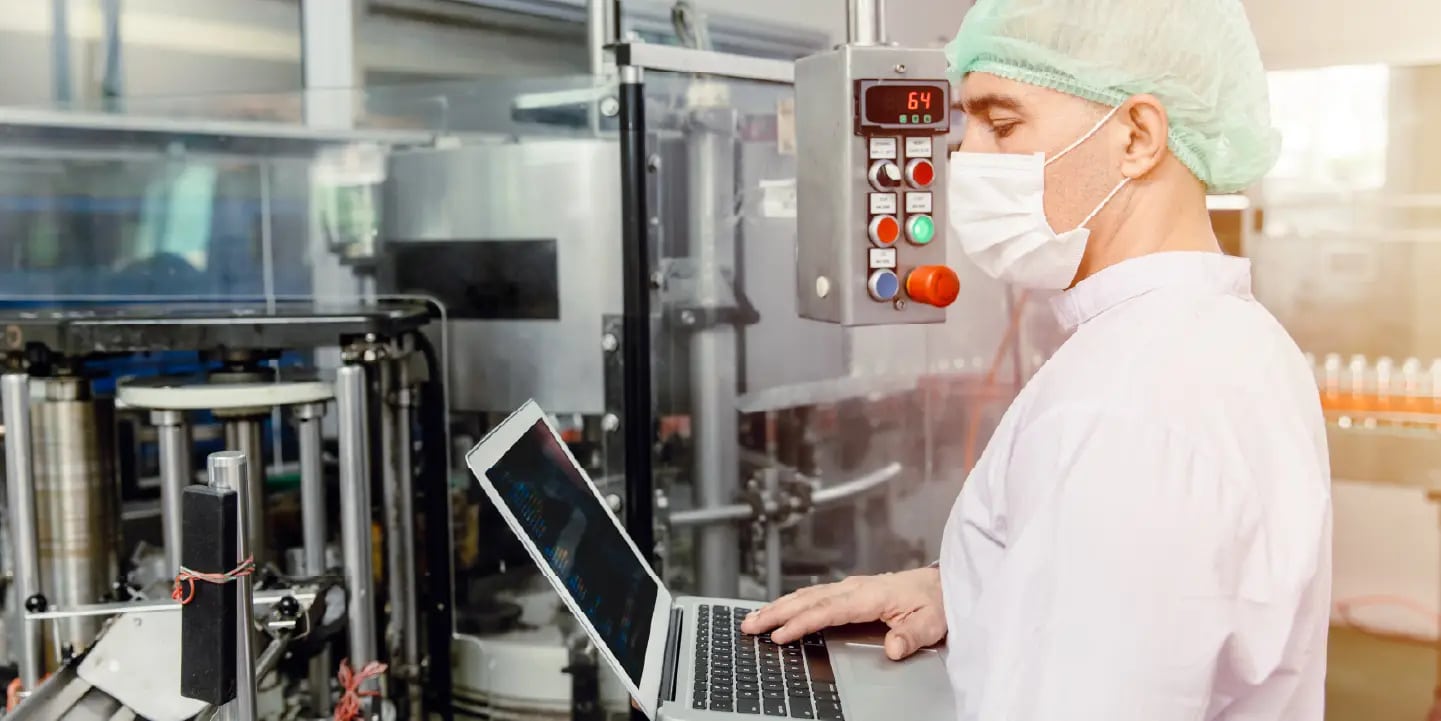 A person in protective clothing and a face mask uses a laptop while monitoring equipment in a factory or laboratory setting.