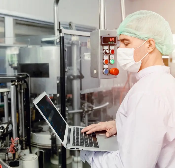 Eine Person in Schutzkleidung und mit Maske arbeitet in einer Fabrik an einem Laptop und steht neben Industrieanlagen und Schalttafeln.