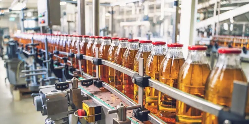 Glass bottles filled with amber liquid are lined up on a conveyor belt in a beverage production facility.