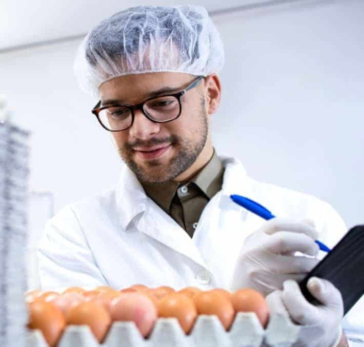 A person in a lab coat, hair net, and gloves inspects a tray of eggs, holding a pen and clipboard, in a laboratory or food inspection setting.