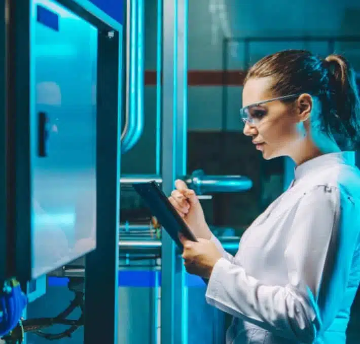 Eine Frau in einem Laborkittel und mit Schutzbrille bedient ein Tablet, während sie neben Industrieanlagen in einem Labor oder einer Fabrik steht.