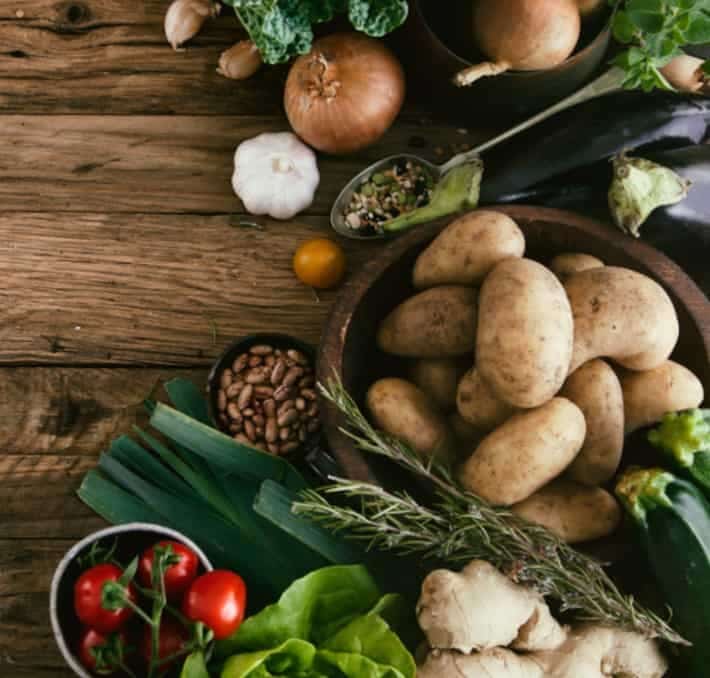 A variety of fresh vegetables including potatoes, tomatoes, zucchini, eggplant, onions, garlic, and leafy greens arranged on a rustic wooden table.
