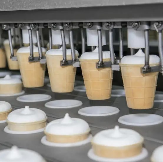 Ice cream cones being filled by an automated machine on a production line, with completed cones and lids visible on the conveyor belt.