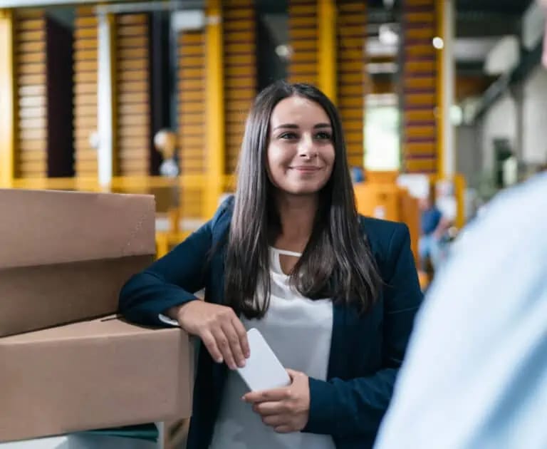 A woman in business attire stands indoors beside stacked cardboard boxes, holding a smartphone and smiling at someone out of frame.