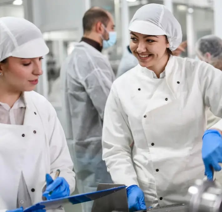 Two workers in white uniforms and hairnets discuss paperwork in a food processing facility, with other staff visible in the background.