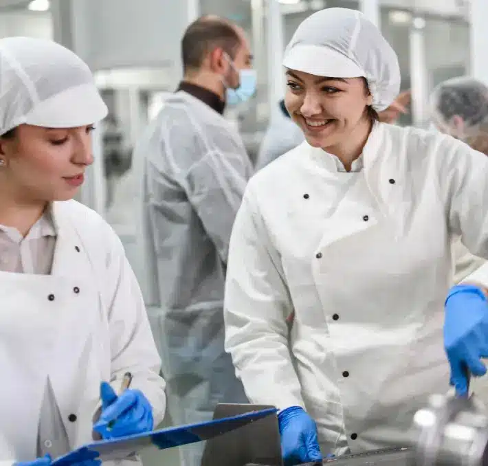 Zwei Mitarbeiter in weißen Uniformen und Haarnetzen besprechen in einem Lebensmittelverarbeitungsbetrieb Verwaltungsangelegenheiten, während im Hintergrund weitere Mitarbeiter zu sehen sind.
