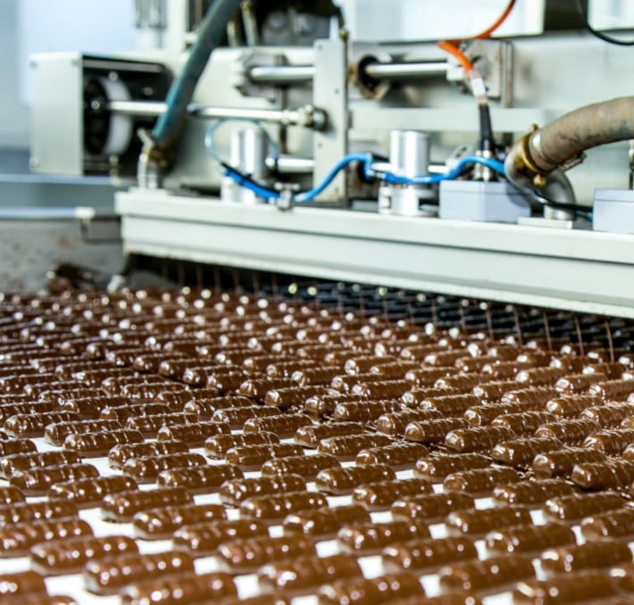 A conveyor belt carries rows of chocolate candies passing through an industrial machine in a factory setting.