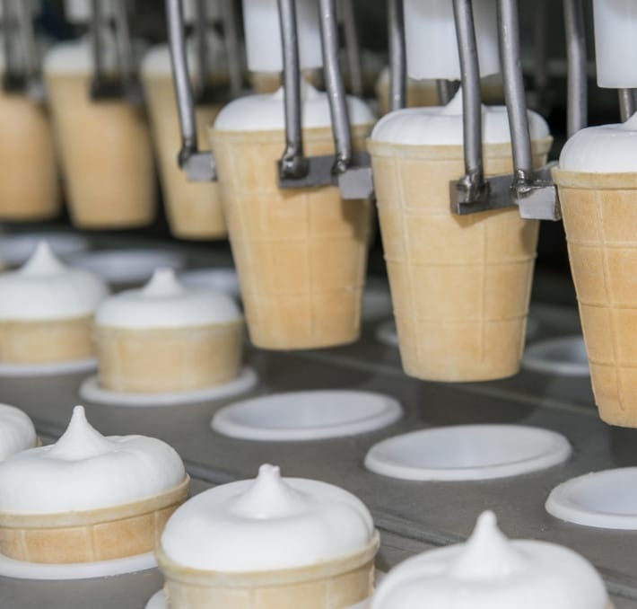 Ice cream cones being filled with vanilla soft serve by a machine in a factory production line.