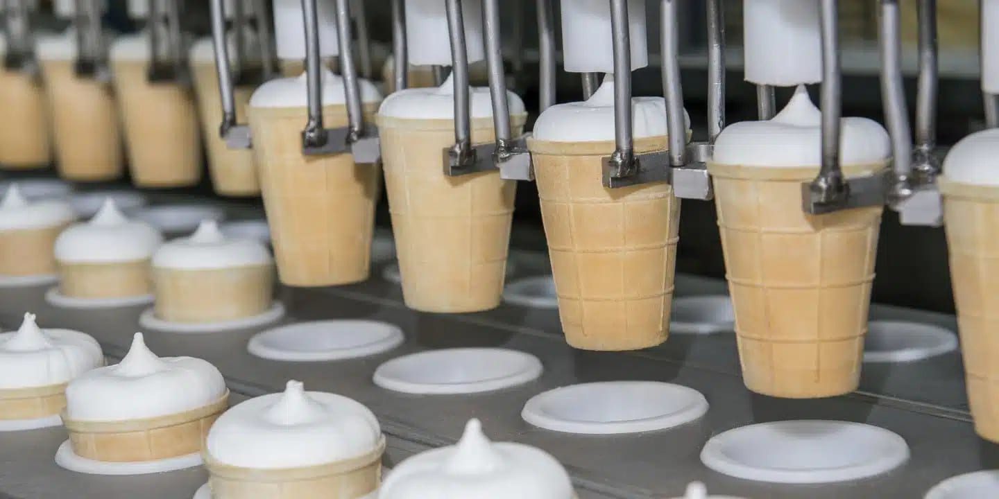 Eistüten, die von einem Automaten auf einer Produktionslinie mit Softeis gefüllt werden, wobei weitere gefüllte Tüten auf einem Förderband zu sehen sind.