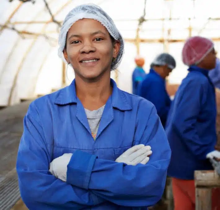 Person wearing a blue lab coat, hair net, and gloves stands with arms crossed in a greenhouse or industrial setting, with others working in the background.
