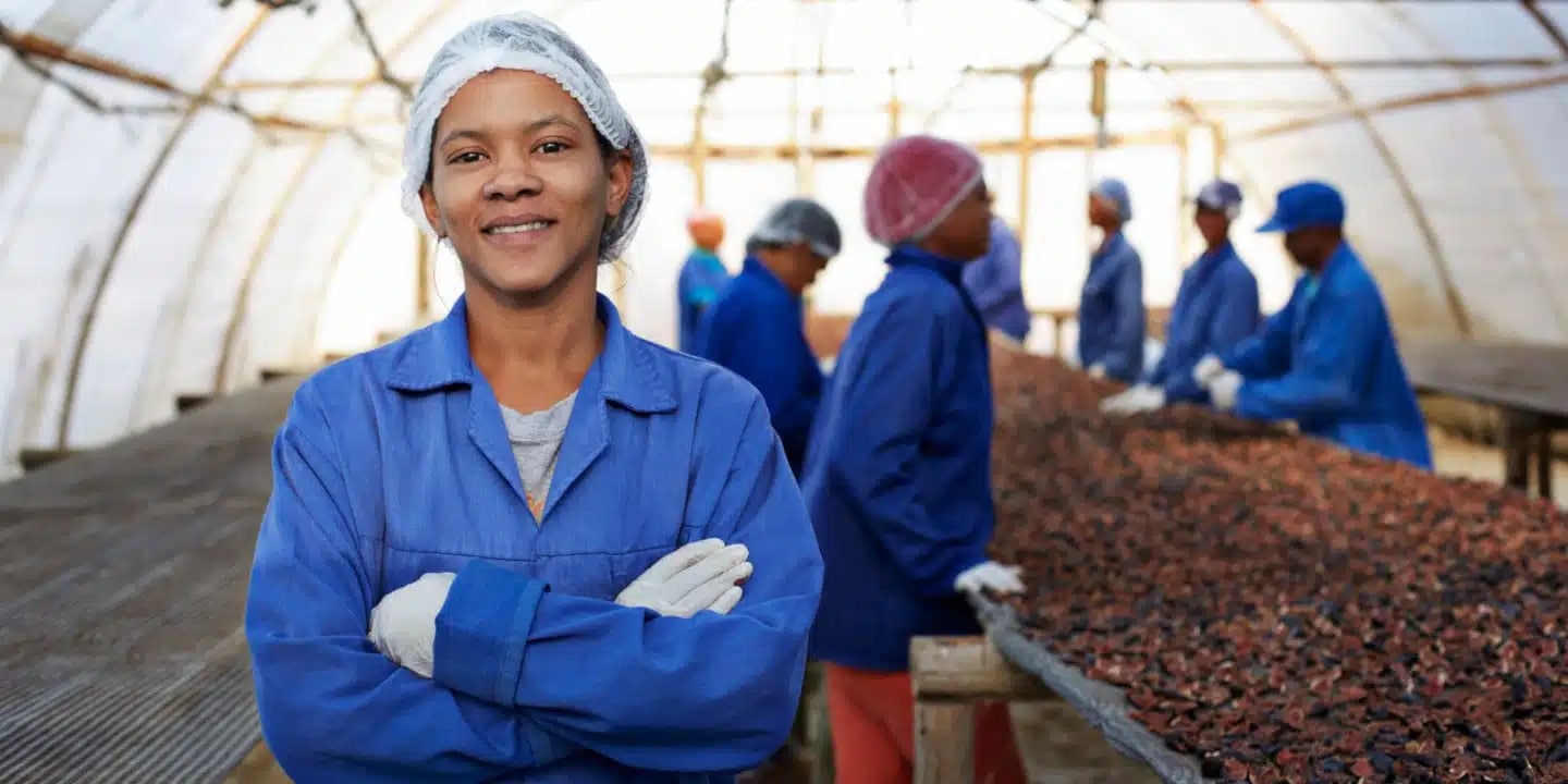A woman in a blue coat and hairnet stands with arms crossed in a greenhouse, with workers sorting dried goods on tables in the background.