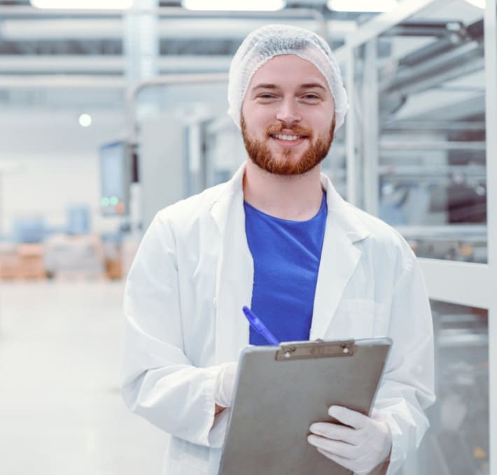 A man in a lab coat, hair net, and gloves holds a clipboard and pen, standing in a clean, industrial or laboratory setting.