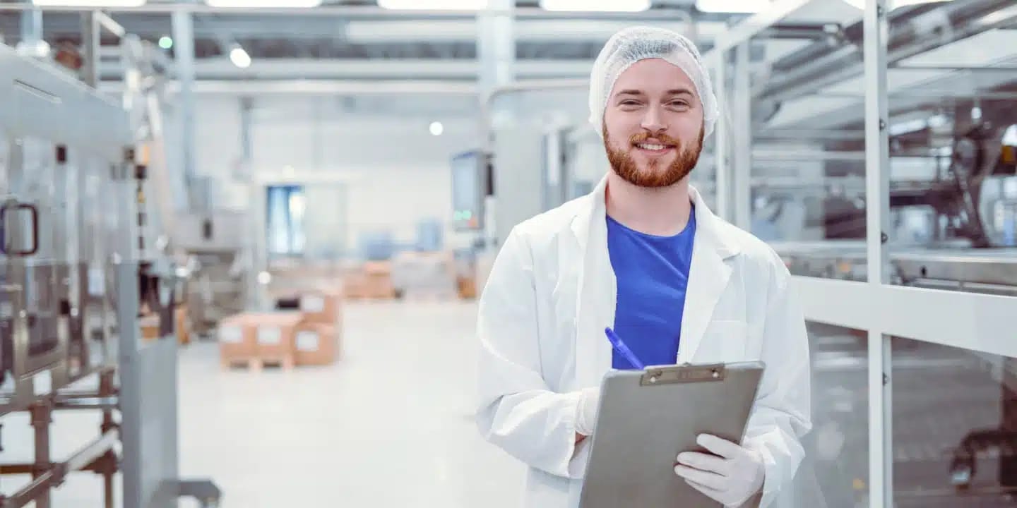 Ein Mann in Laborkittel, Haarnetz und Handschuhen, der ein Klemmbrett und einen Stift in der Hand hält und in einer sauberen, modernen Industrie- oder Laborumgebung steht.