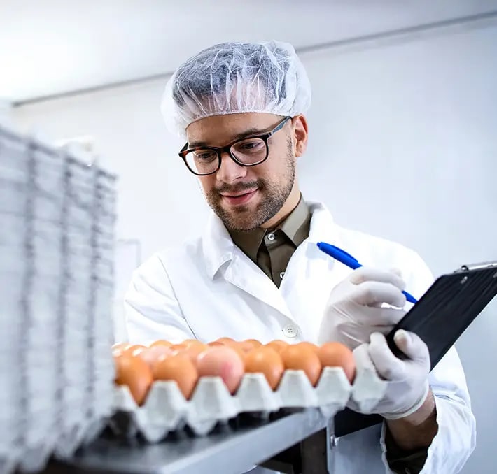 A man in a lab coat, hair net, and gloves inspects eggs in trays and writes notes on a clipboard in a laboratory or food inspection setting.