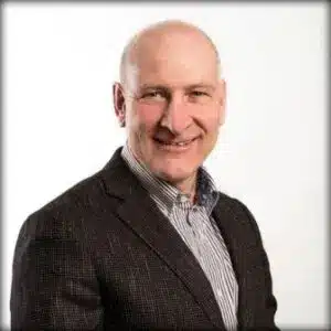 A middle-aged man wearing a dark blazer and striped shirt poses against a plain white background, smiling at the camera.