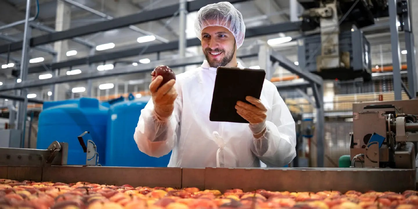 Eine Person in Schutzkleidung inspiziert ein Stück Obst und hält ein digitales Tablet in einer Lebensmittelverarbeitungsanlage.