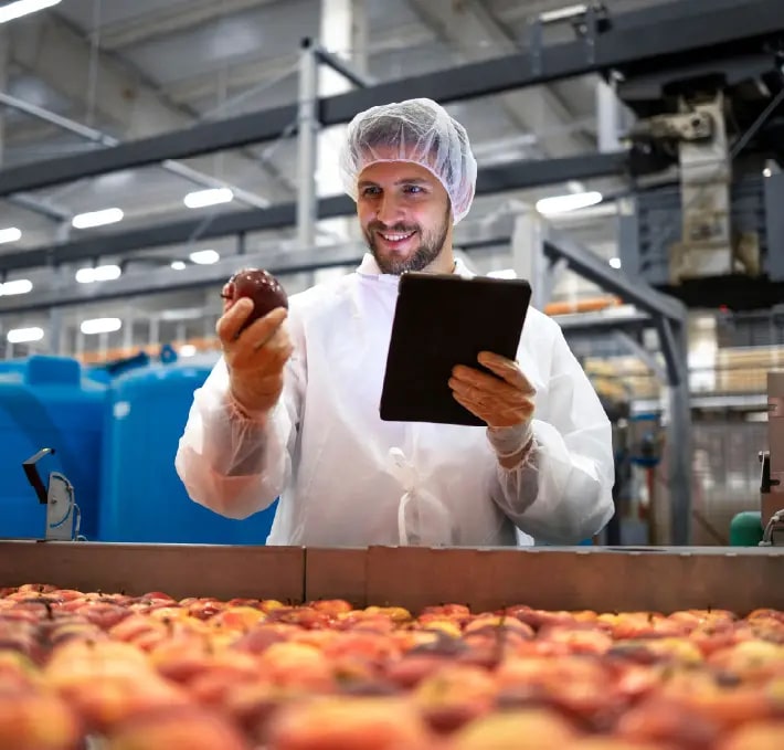 A worker in protective clothing inspects an apple and holds a tablet in a fruit processing facility.