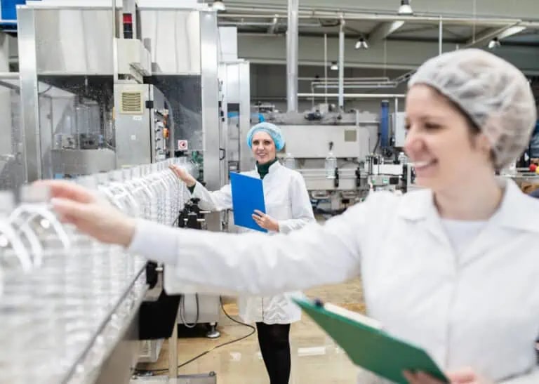 Zwei Arbeiter in weißen Laborkitteln und Haarnetzen kontrollieren Plastikflaschen an einem Fließband in einer Fabrik, halten Klemmbretter in der Hand und lächeln.