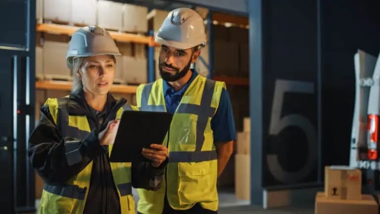 Two workers wearing safety vests and hard hats review information on a tablet in a warehouse or industrial setting.