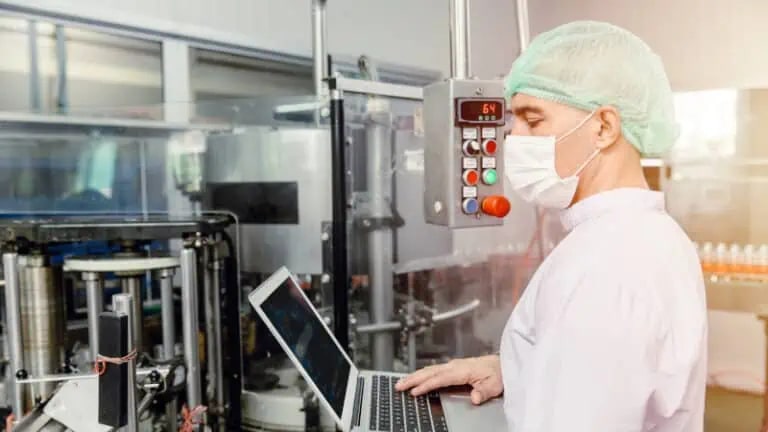 A person in protective clothing and a face mask uses a laptop while standing next to industrial machinery in a factory setting.