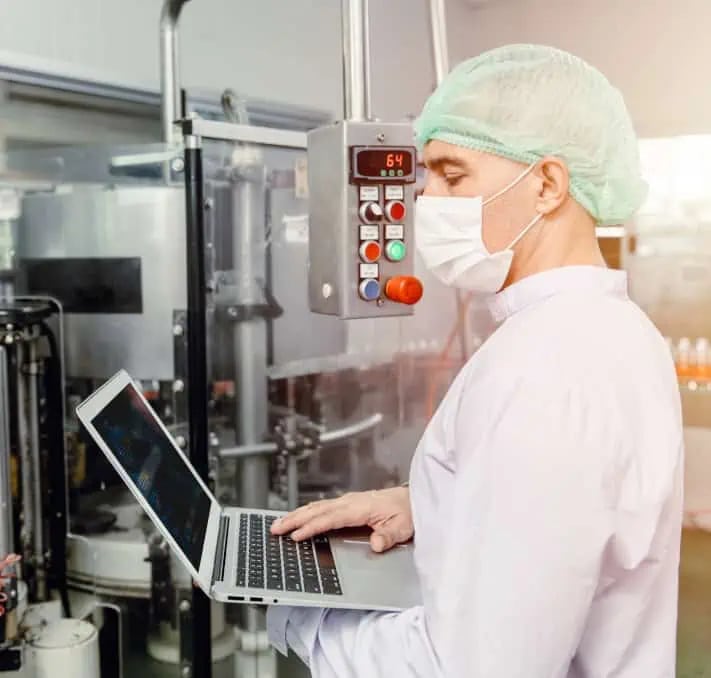 Eine Person in Schutzkleidung, Maske und Haarnetz arbeitet mit einem Laptop, während sie in einer Fabrikanlage industrielle Geräte überwacht.