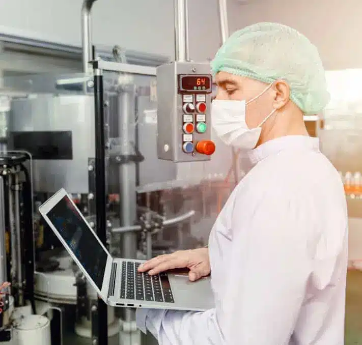 A person in protective clothing, mask, and hairnet uses a laptop while monitoring industrial equipment in a factory setting.