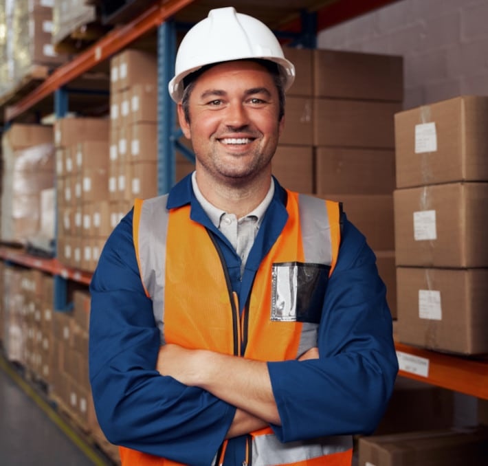 A warehouse worker wearing a white hard hat and orange safety vest stands smiling with arms crossed in front of shelves stacked with cardboard boxes.