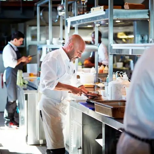 A chef in a white shirt and apron prepares food at a stainless steel counter in a busy commercial kitchen, with other staff working in the background.